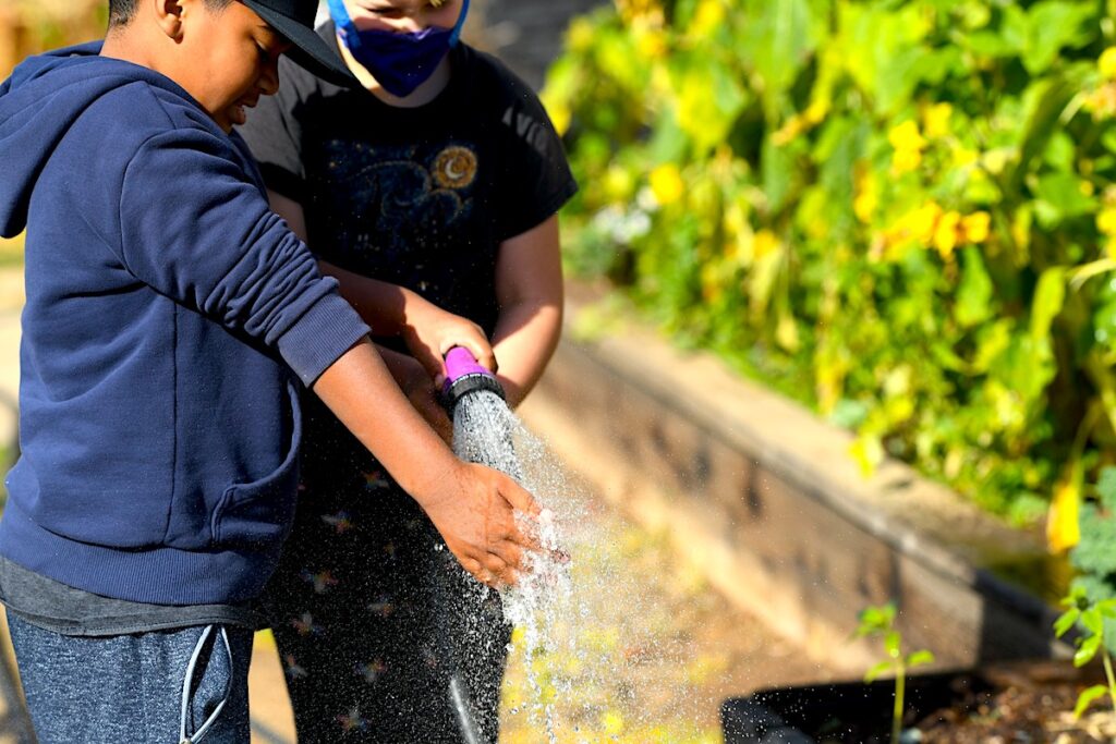 Two students at San Diego Cooperative Charter School hold a water hose next to a school garden